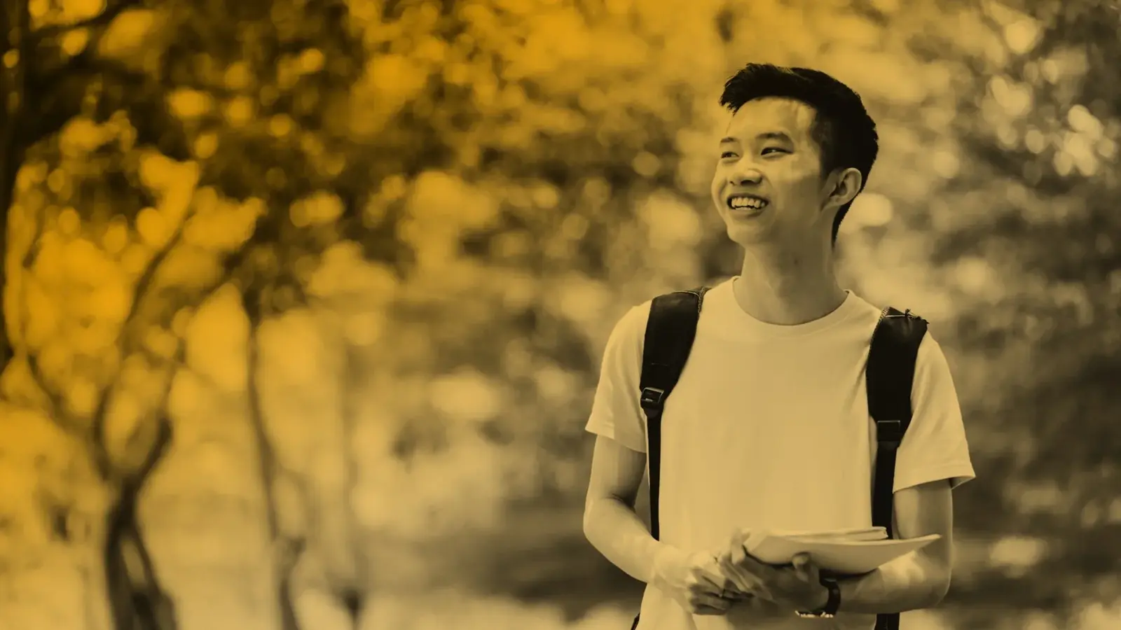 Smiling student with a backpack holding books while walking outdoors among trees