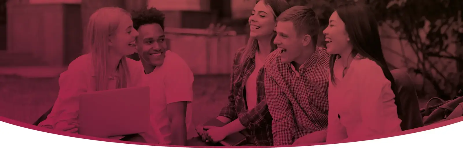 A group of students sitting outdoors together, smiling and talking around an open laptop