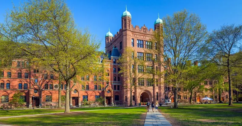 Students walking a path across a sunny green lawn toward a Gothic red-brick university building with green-domed towers.