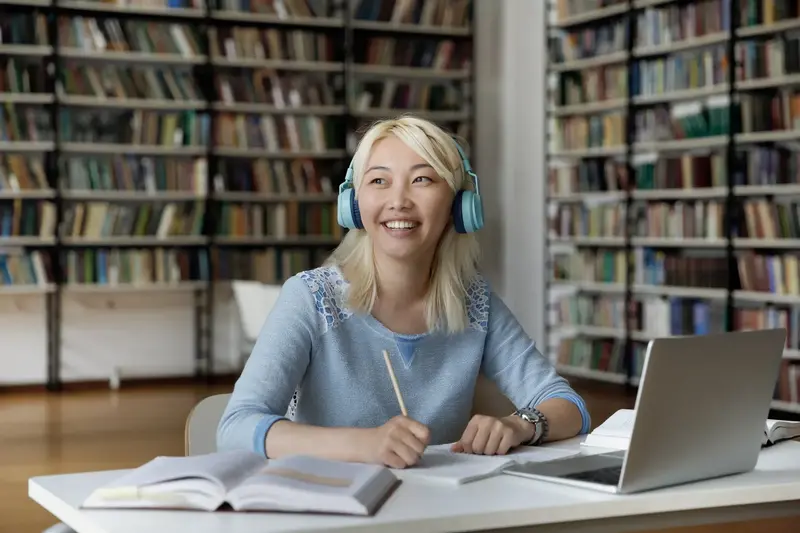 Smiling student wearing blue headphones sits with a pencil, notebook and laptop at a library table lined with bookshelves