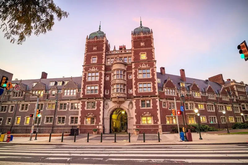 Red brick collegiate Gothic university building with twin towers and an arched entrance at dusk, seen across an empty crosswalk.