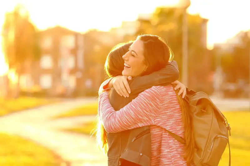 Two smiling students hugging outdoors at sunset, one wearing a backpack
