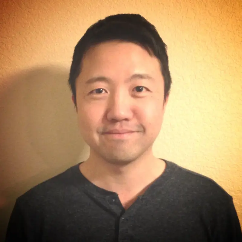 Man in a dark gray henley shirt smiling slightly in front of a warm beige wall