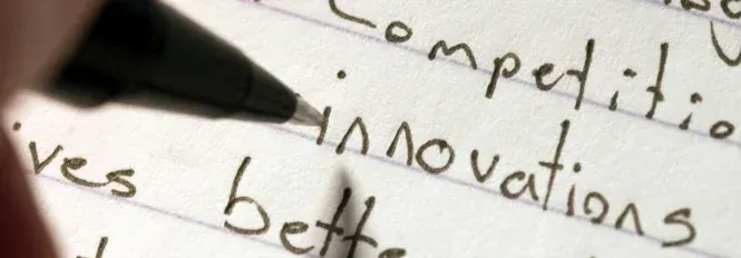 A pen poised over lined paper with handwritten words including "competition," "innovation" and "better".